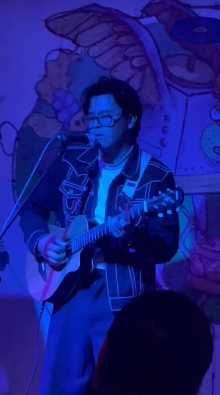 Ciudades Imagen de una presentación musical en vivo donde una persona canta y toca la guitarra en un escenario con iluminación azul.