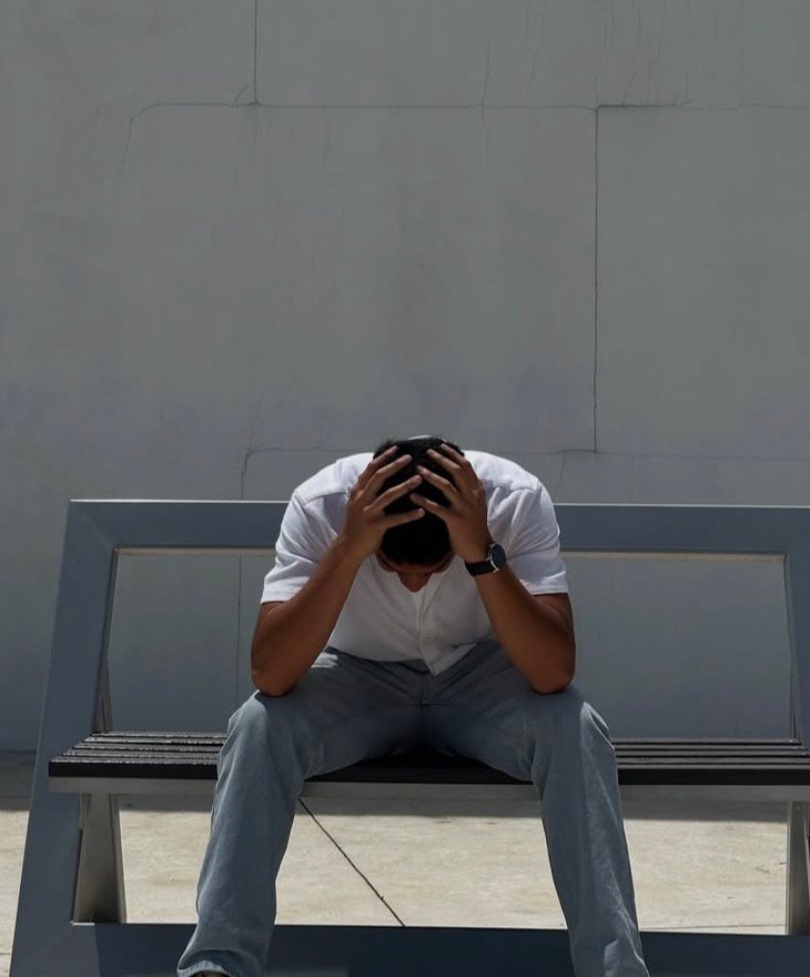 Lanzamientos Hombre sentada en una banca con la cabeza entre las manos frente a una pared gris.